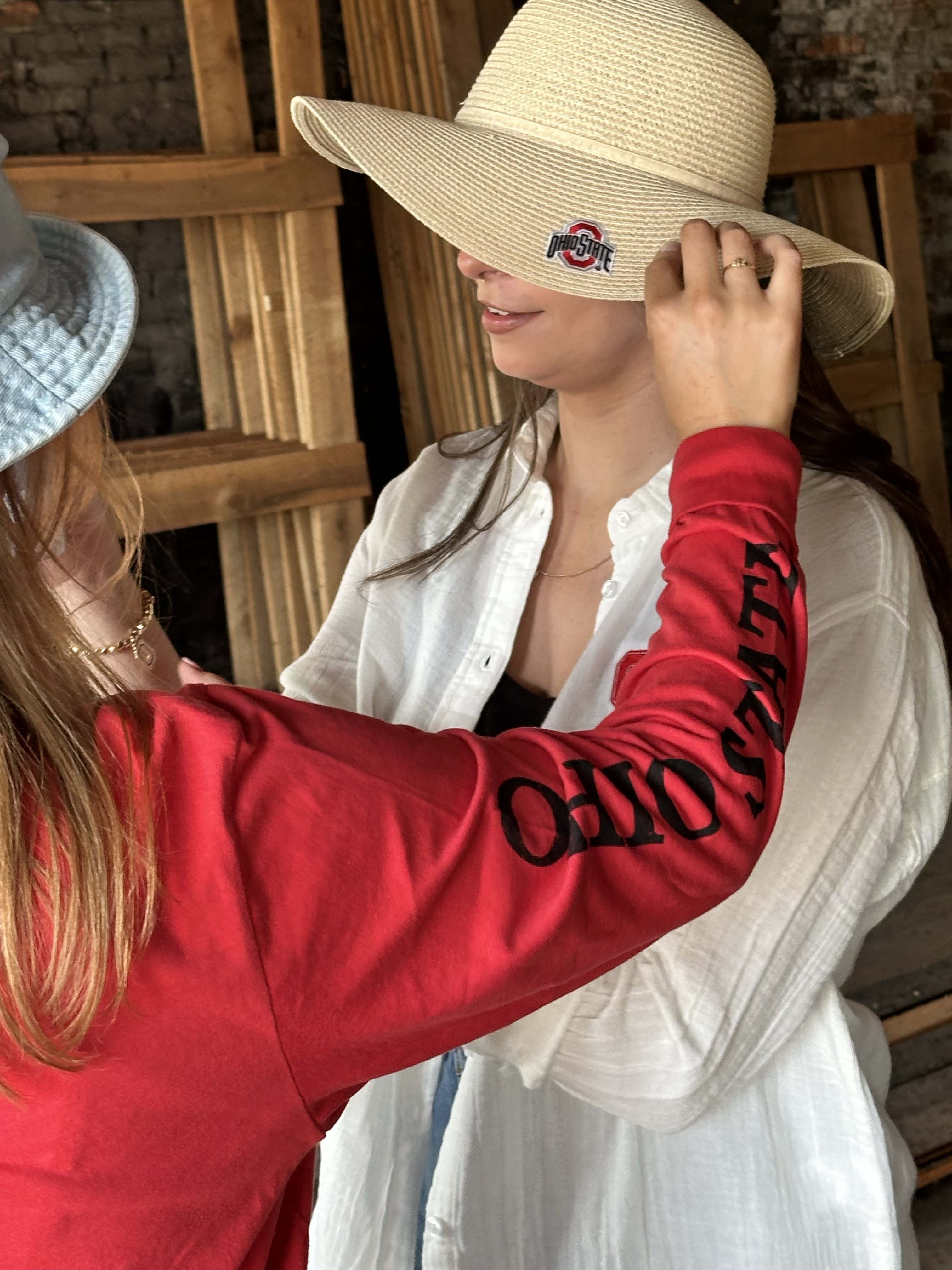 A woman wearing a large beige straw sun hat with a black and red Ohio State Buckeyes logo patch on the brim, while another person in a red sweatshirt adjusts the brim. The background shows a rustic wooden structure.