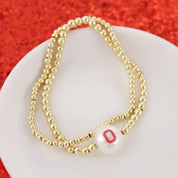 A double-strand beaded necklace with gold-toned beads and a white pearl bead featuring a red letter 'O' charm, displayed on a white plate against a textured red background.