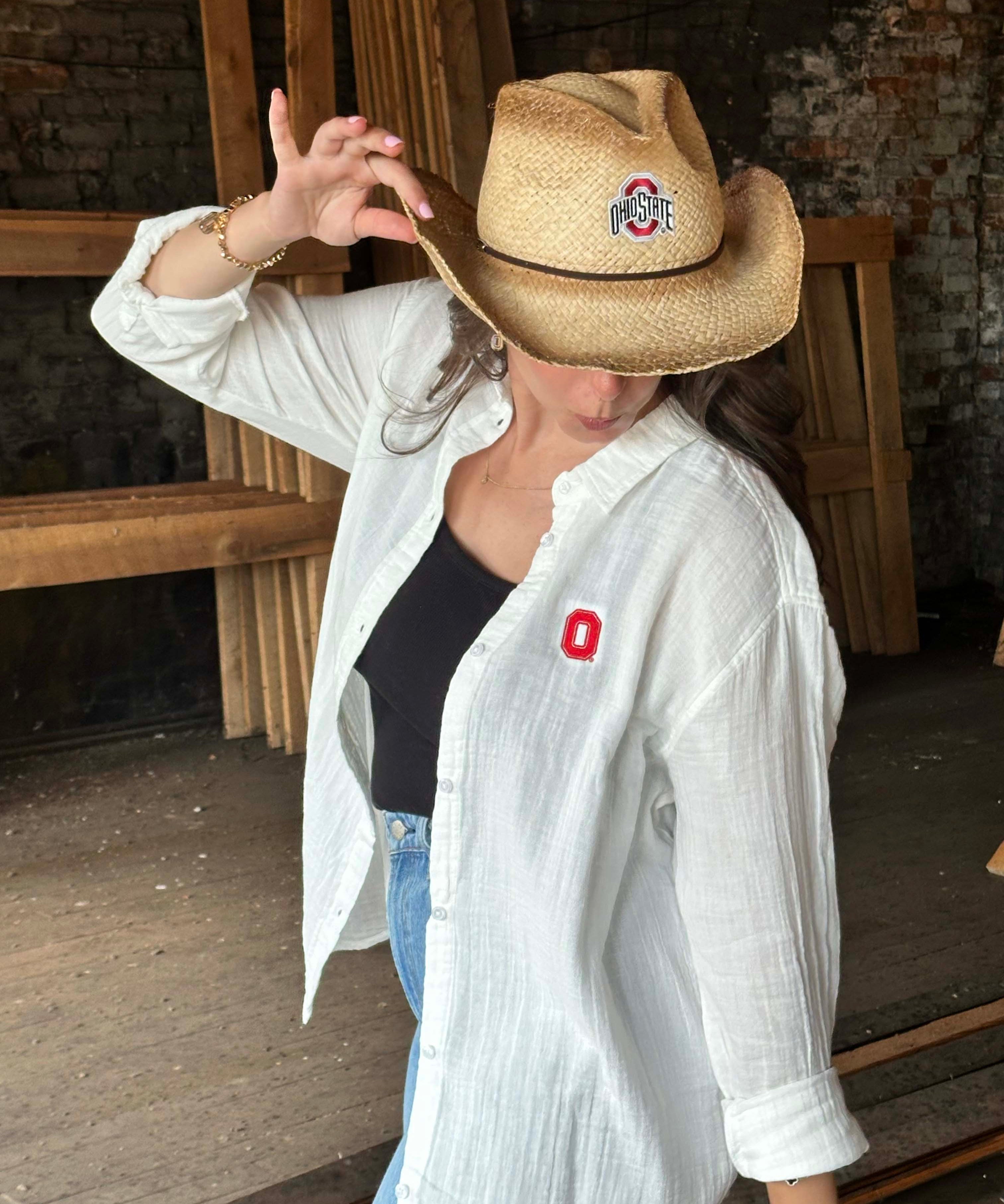 A woman wearing a straw cowboy hat with an Ohio State Buckeyes logo patch and a white button-down shirt with a red Ohio State 'O' logo on the chest, posing in a rustic indoor setting.