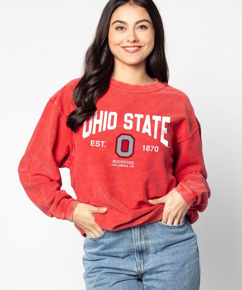 A woman modeling a red washed sweatshirt with 'OHIO STATE' and the Buckeye 'O' logo printed in white on the front, paired with light blue jeans against a light gray studio background.