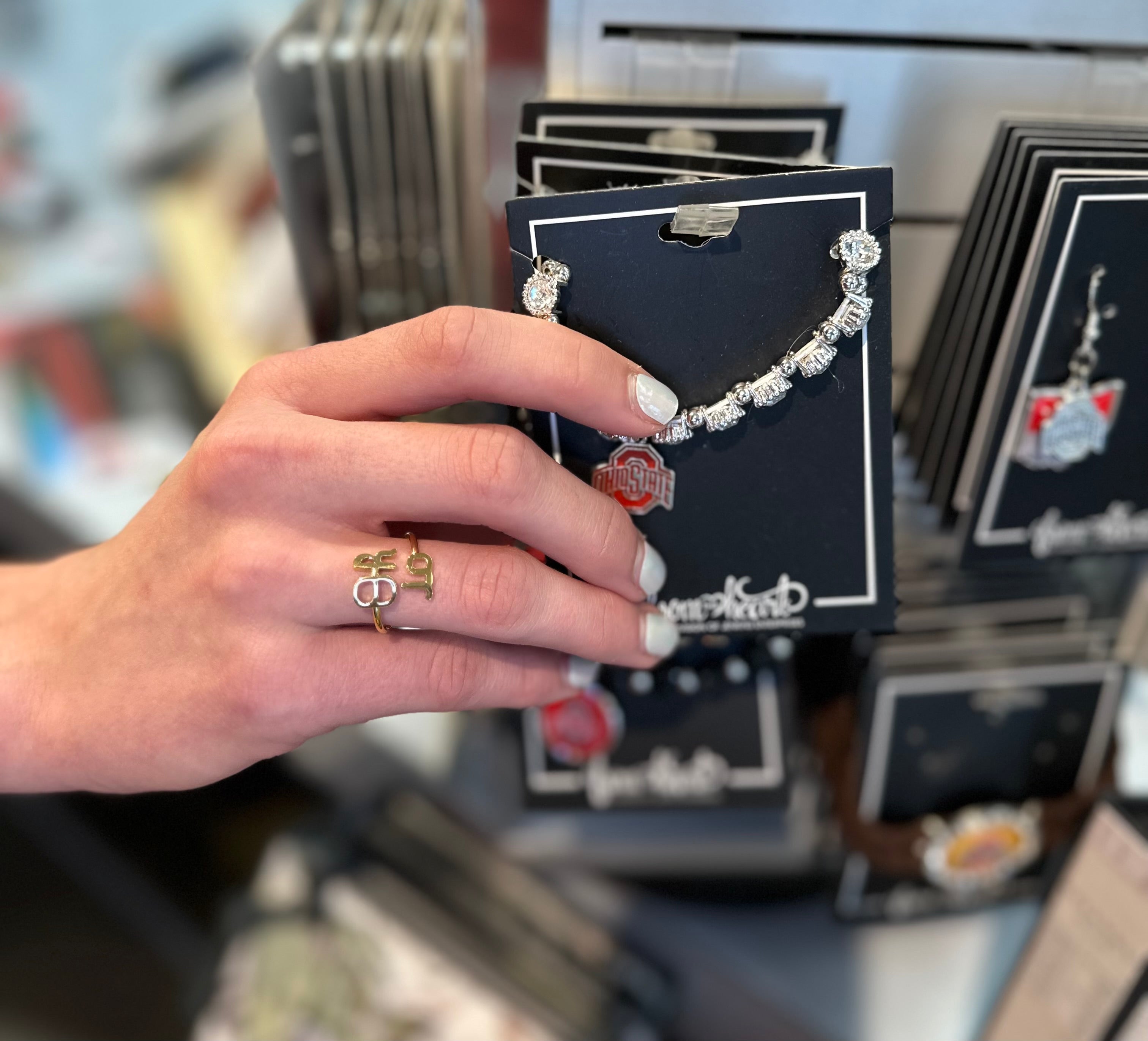 A hand with white nail polish holds a silver beaded bracelet with a red and white Ohio State Buckeyes logo charm on a black display card. The background shows a retail store display with other jewelry cards.