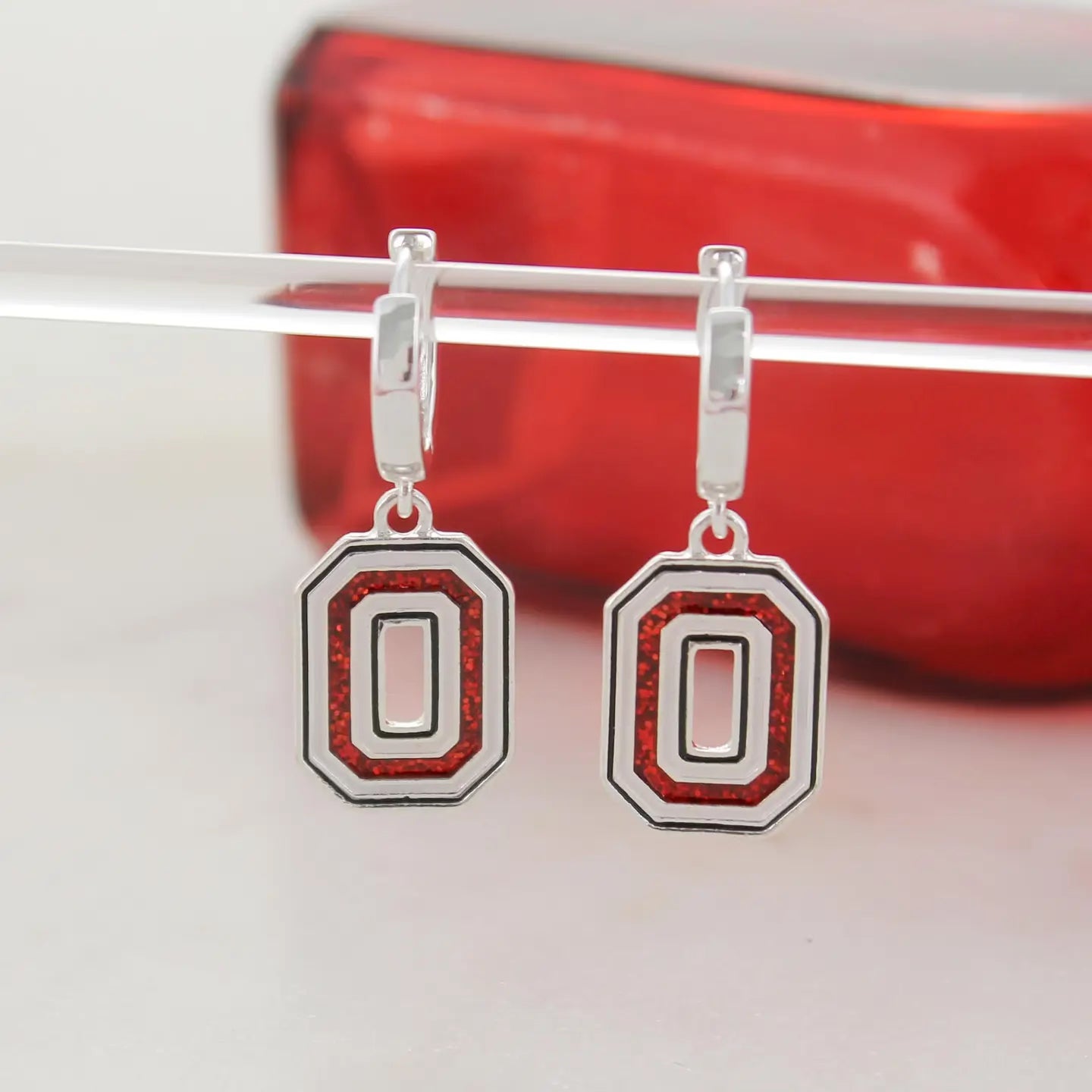 A pair of silver drop earrings featuring a large letter 'O' design with red glitter-style lettering, hanging from a clear display stand against a red and white background.
