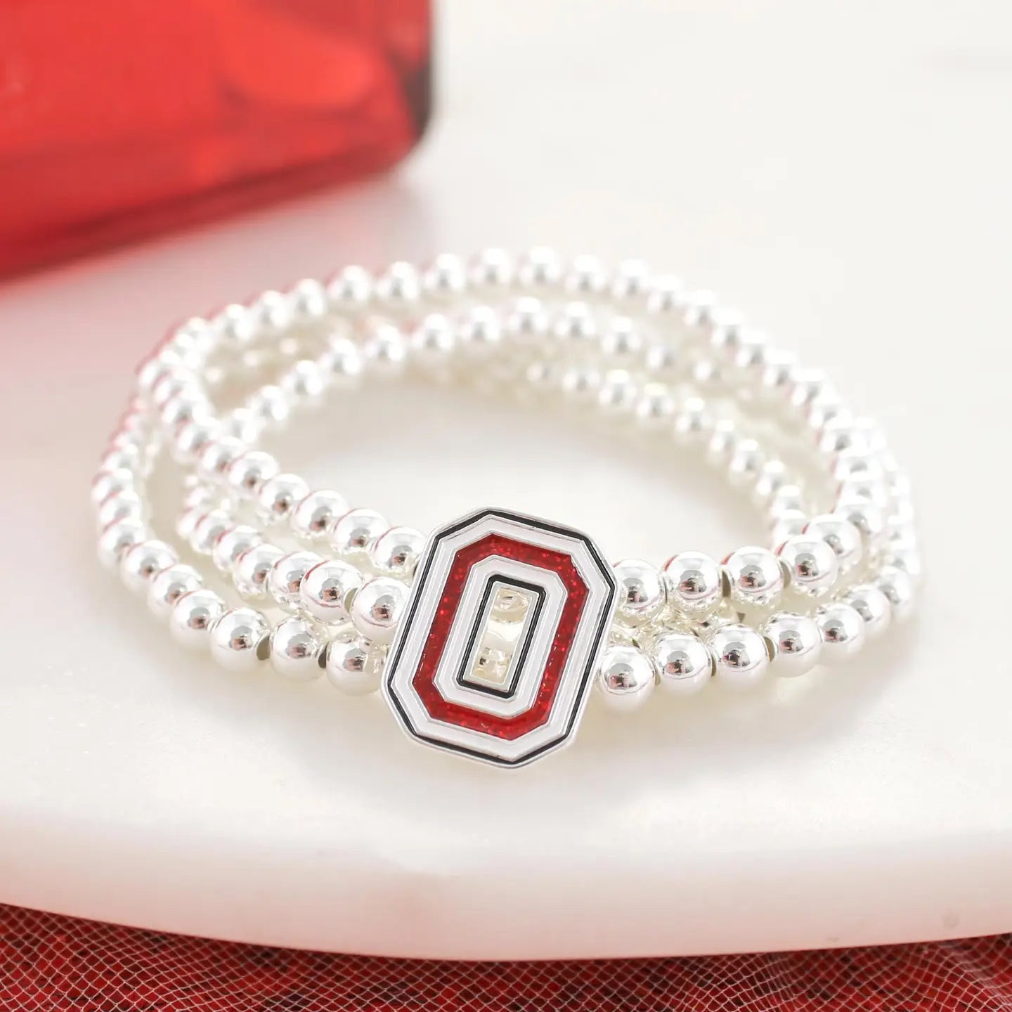 A close-up of a multi-strand silver beaded bracelet with a central octagonal charm featuring red, white, and black concentric rings, resting on a white surface.