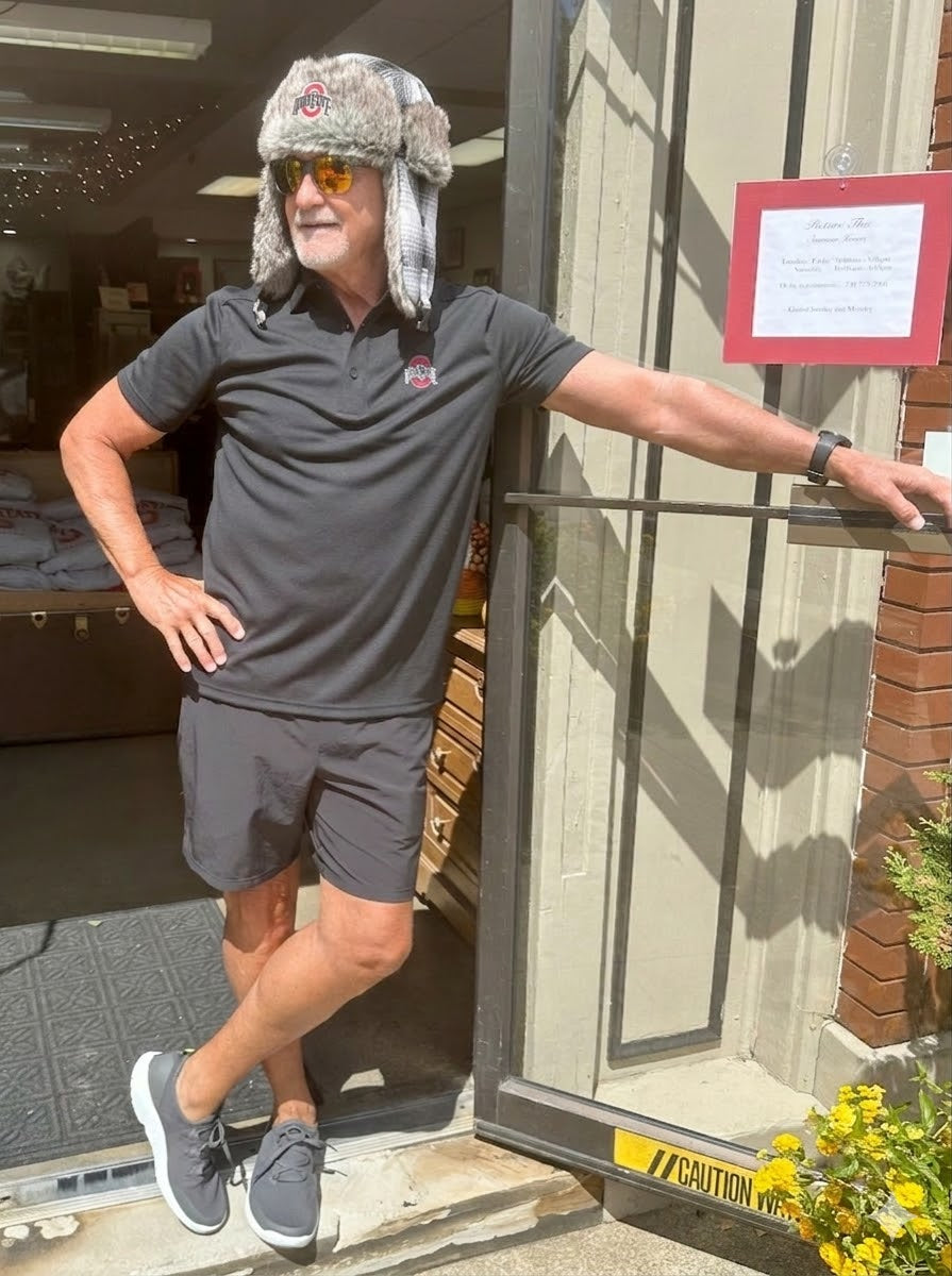 Man wearing a gray shirt and shorts with a fur hat, standing outside a building.