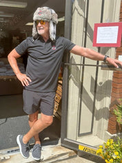 Man wearing a gray shirt and shorts with a fur hat, standing outside a building.