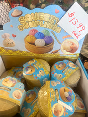 A retail display box filled with small round plastic toy dumpling containers. The top card reads 'SQUEEZI DUMPLINGS' with cartoon dumplings and a hand squeezing one, plus a handwritten price tag showing $13.