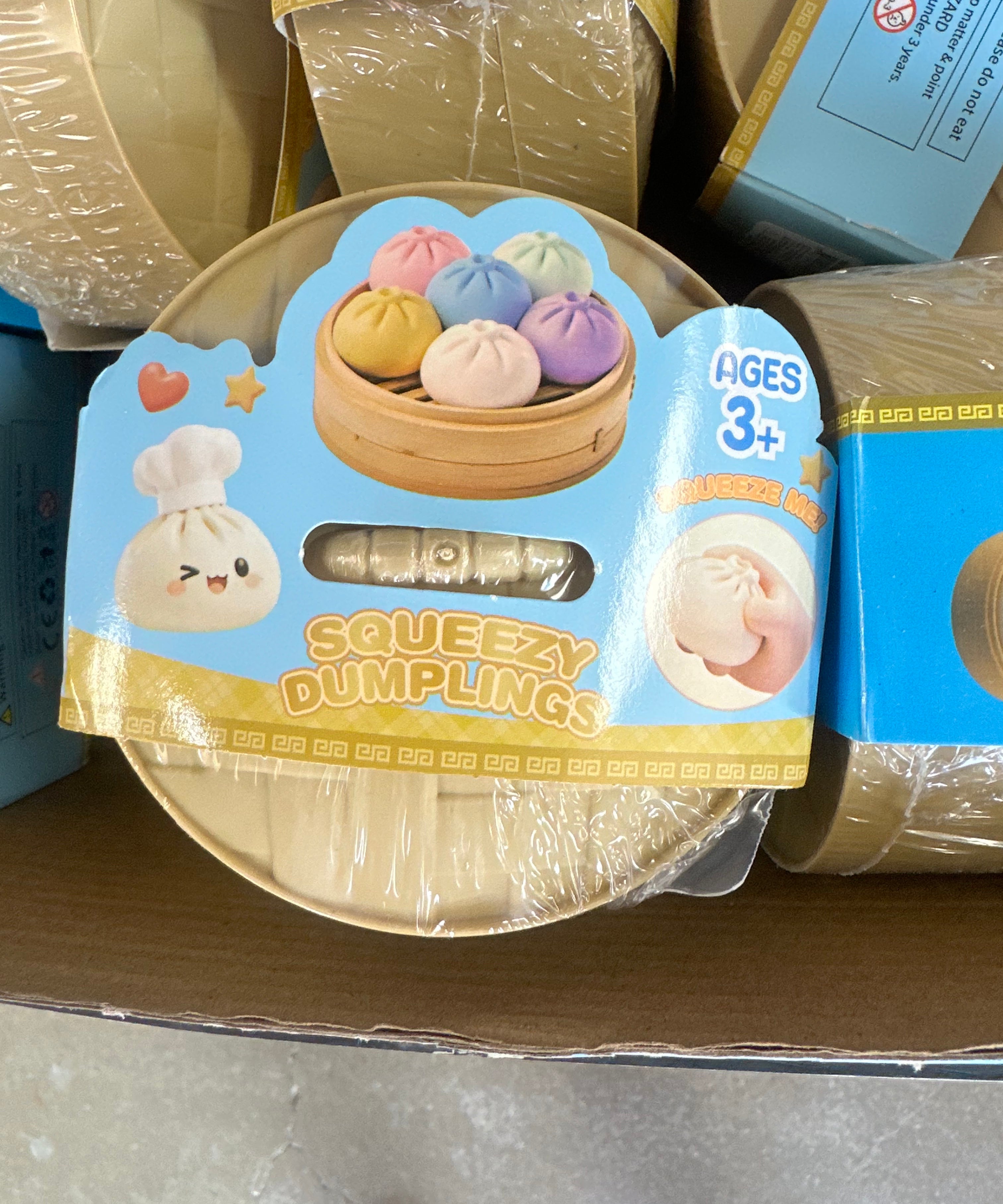 A round toy container labeled 'Squeezzy Dumplings' with a cartoon dumpling character and a photo of colorful dumplings in a steamer, shown on a cardboard shelf next to other similar packages.