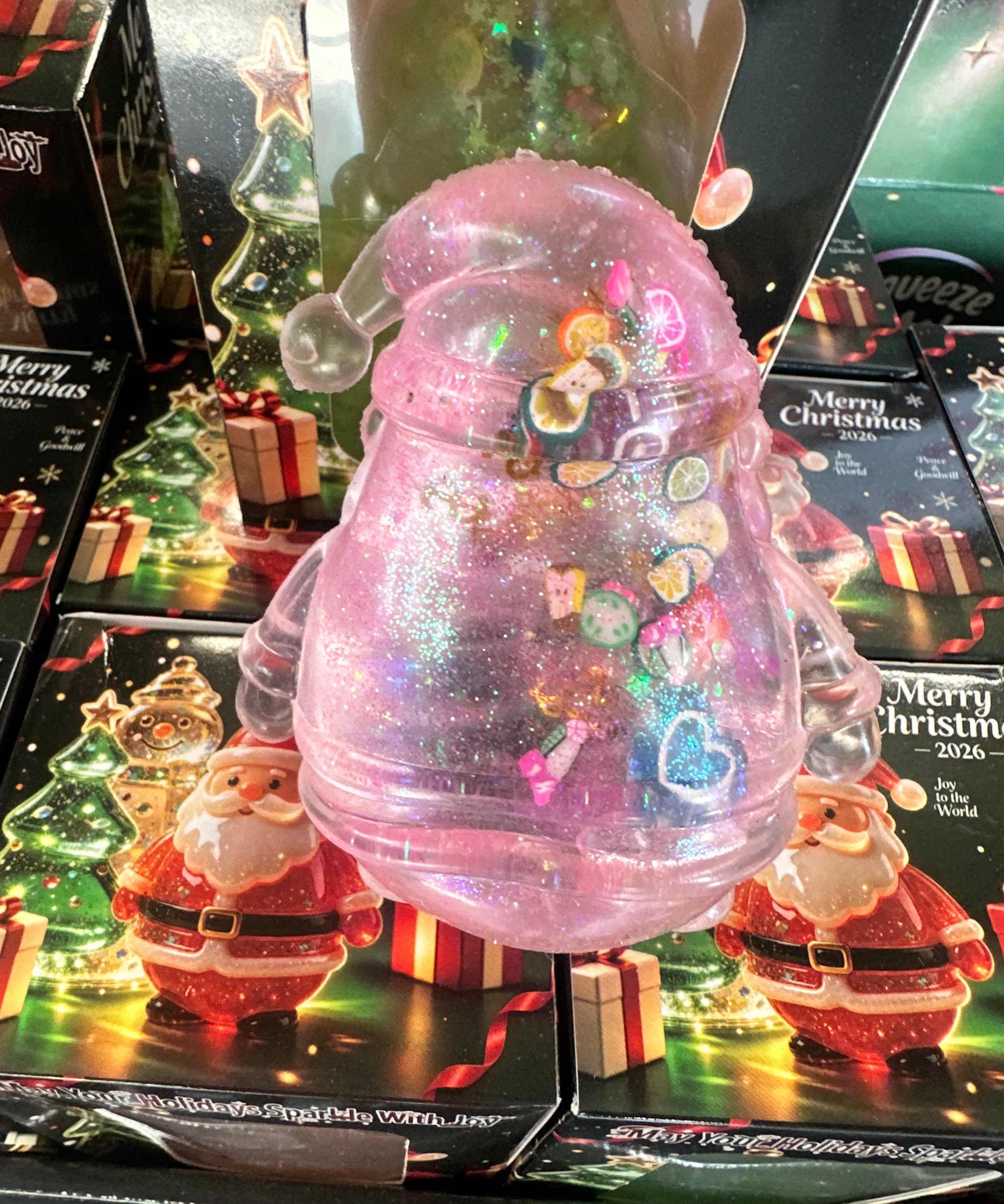 A pink glittery Christmas ornament-shaped container with a clear Santa hat and colorful heart-shaped embellishments, displayed in front of holiday-themed packaging featuring Santa Claus and 'Merry Christmas' designs.