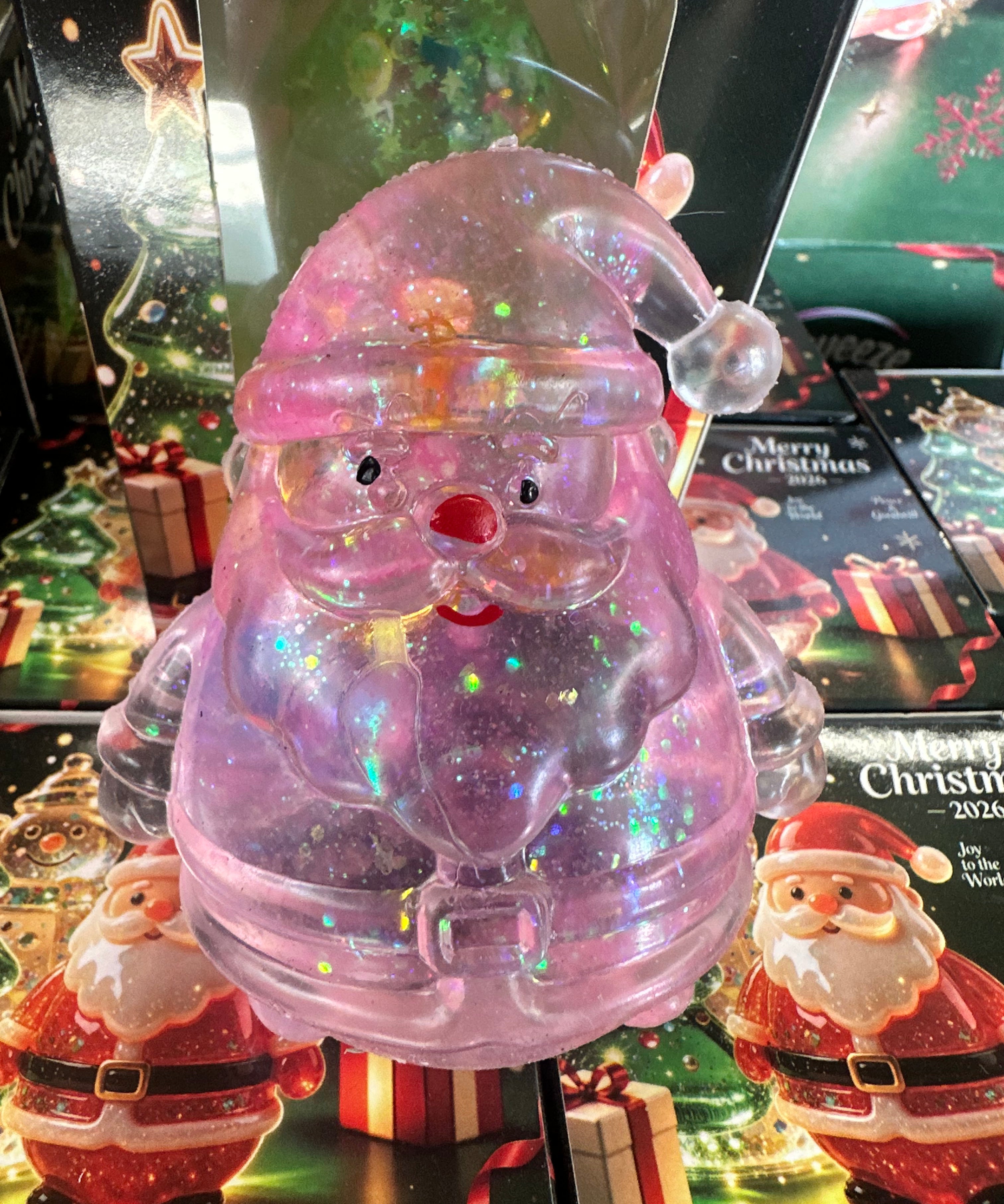 A translucent pink Santa Claus figurine filled with iridescent glitter, displayed in front of Christmas-themed packaging and other Santa decorations.
