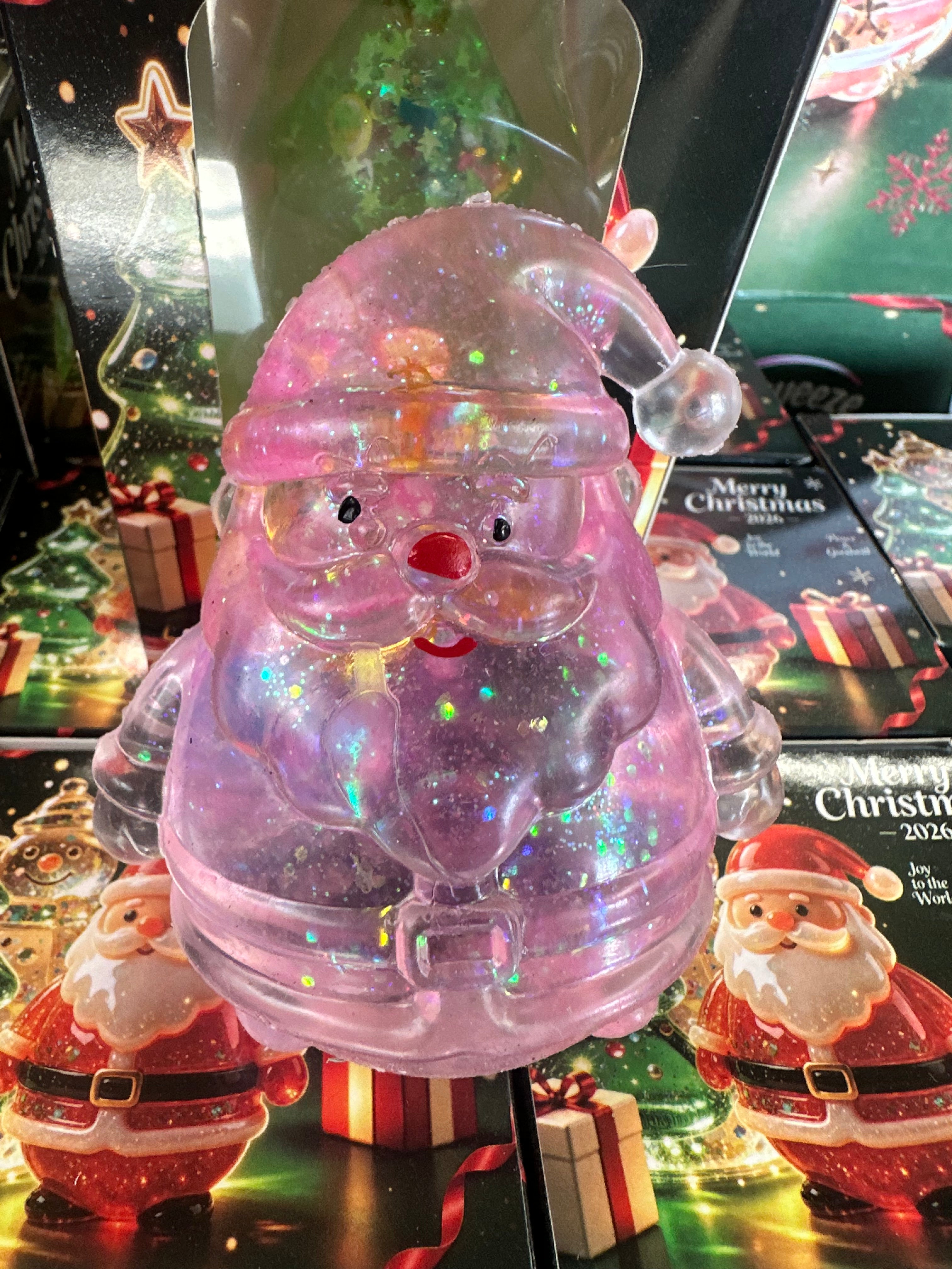 A translucent pink Santa Claus figurine filled with iridescent glitter, displayed in front of Christmas-themed packaging and other Santa decorations.