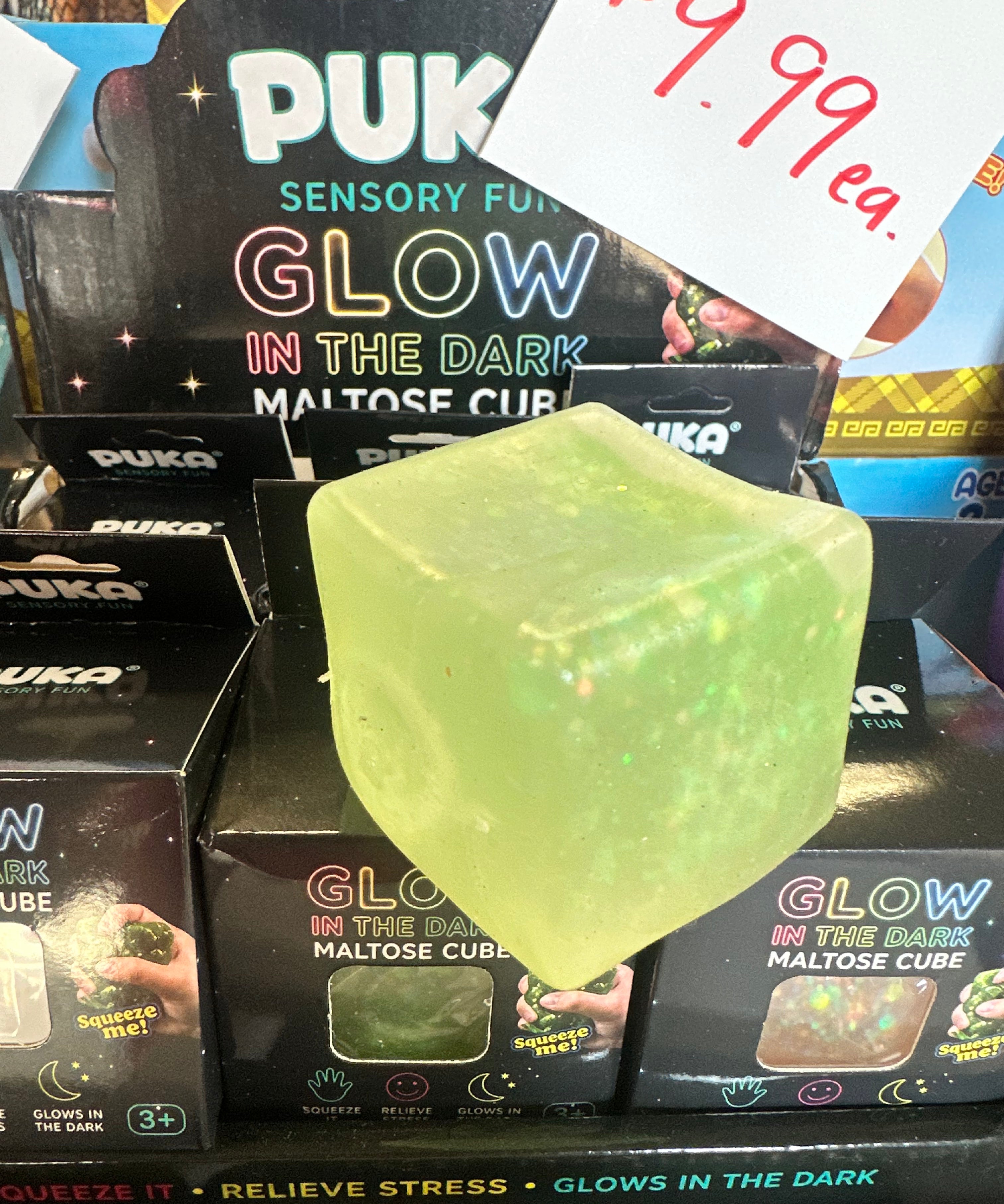 A translucent pale green glow-in-the-dark maltose cube toy with embedded glitter, displayed in front of its retail packaging. A handwritten price tag reading '$9.