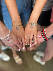 Several people standing together with their hands stacked in the center, wearing silver and gold bracelets, rings, and a small red and white Ohio State Buckeyes logo on one wrist.