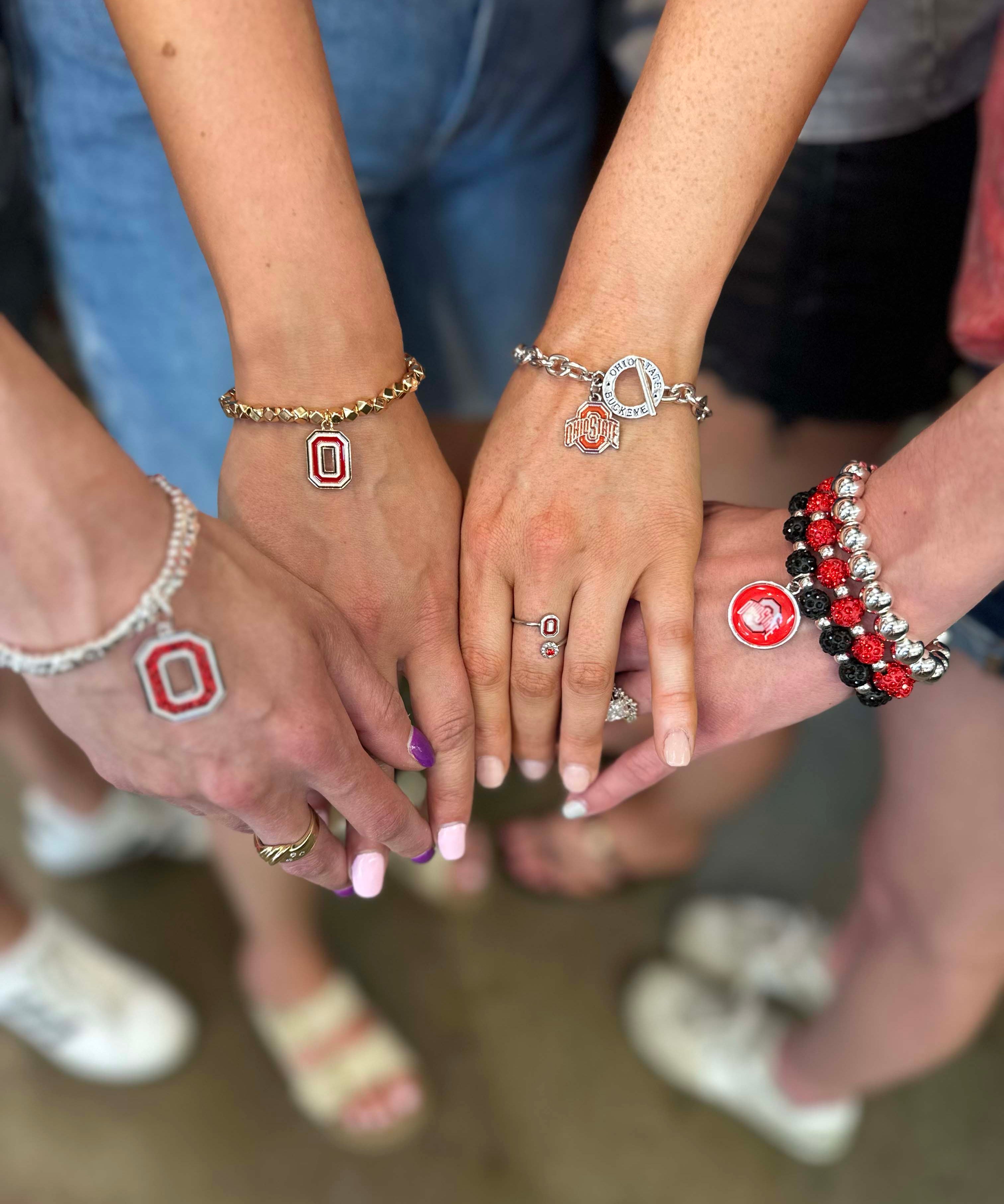 Several people standing together with their hands stacked in the center, wearing silver and gold bracelets, rings, and a small red and white Ohio State Buckeyes logo on one wrist.