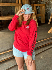 A woman wearing a bright red long-sleeve Ohio State Buckeyes sweatshirt, light blue denim shorts, and a light blue baseball cap with the Ohio State logo, standing in a rustic wood-paneled room.