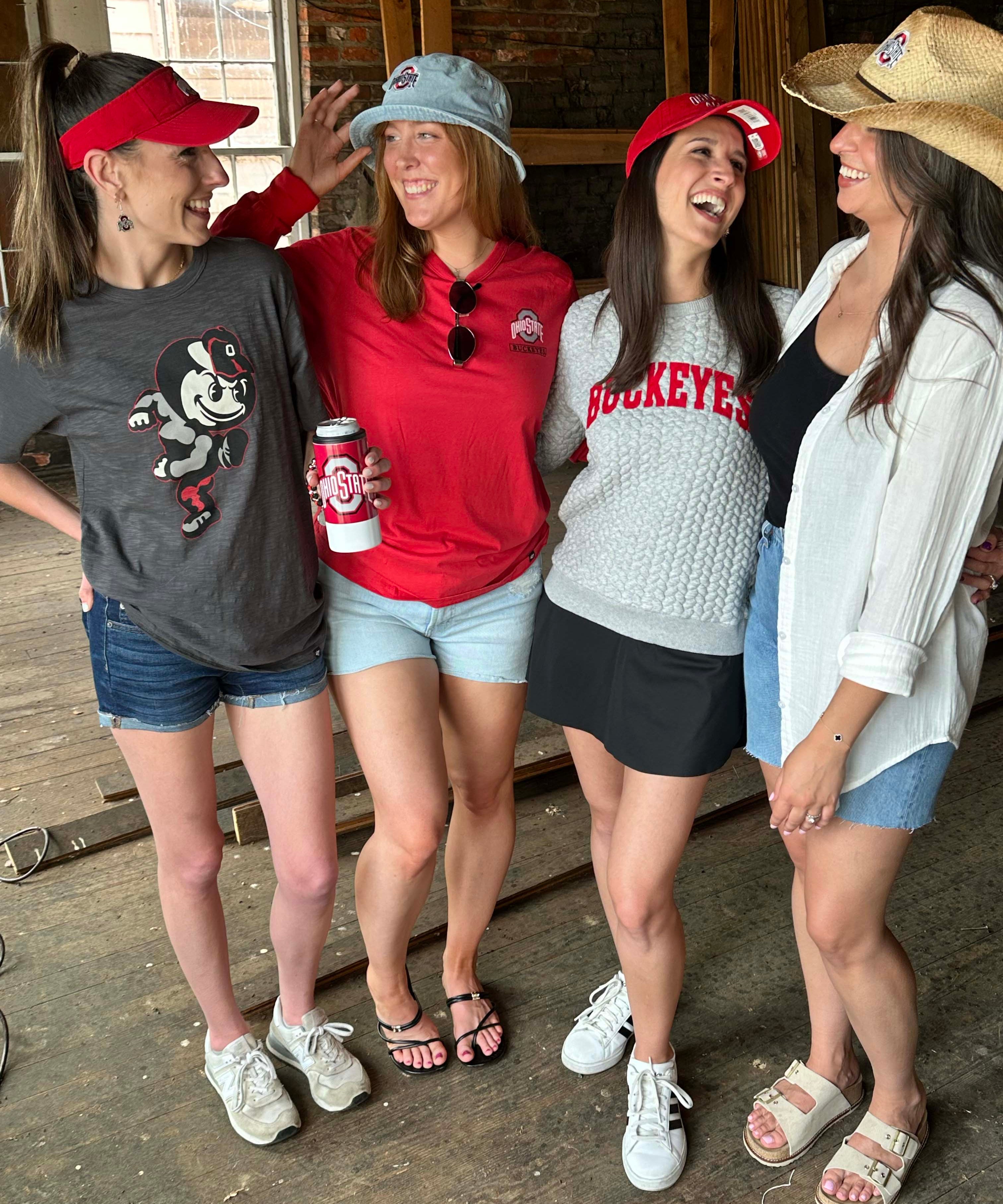 Four women laughing together in a rustic indoor setting, wearing Ohio State Buckeyes themed apparel including a gray graphic t-shirt, a red long-sleeve shirt, a gray knit 'Buckeyes' sweater, and a white button-up shirt.