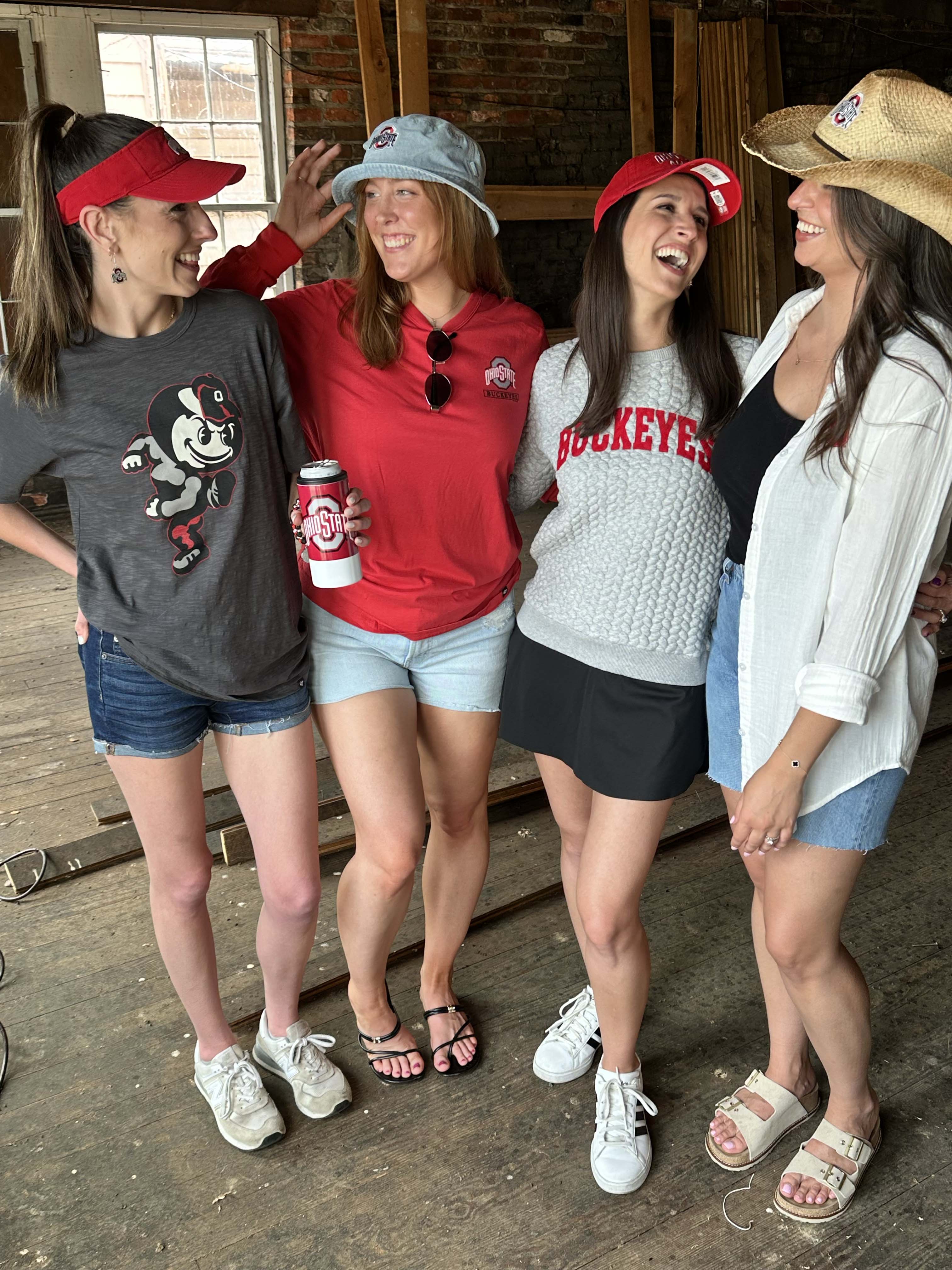 Four women laughing together in a rustic indoor setting, wearing Ohio State Buckeyes themed apparel including a gray graphic t-shirt, a red long-sleeve shirt, a gray knit 'Buckeyes' sweater, and a white button-up shirt.