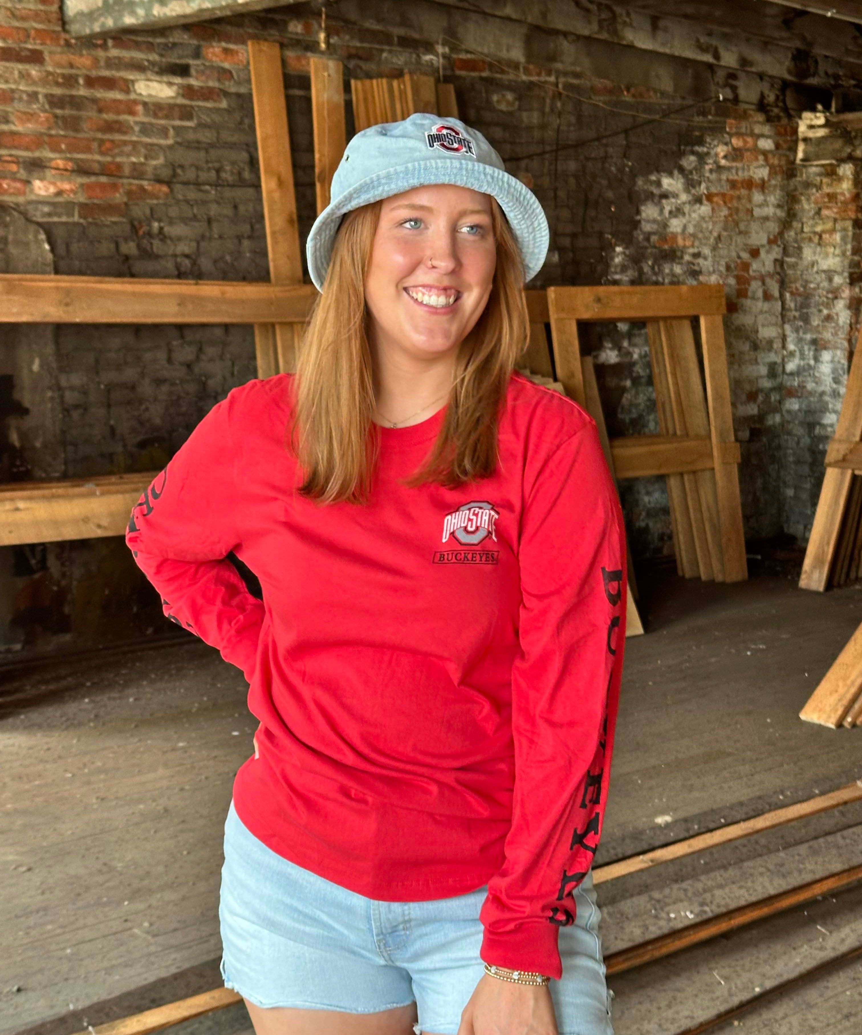 A smiling woman wearing a red long-sleeve t-shirt with a 'Pittsford' logo, a light blue bucket hat, and denim shorts, standing in a rustic workshop with brick and wood walls.
