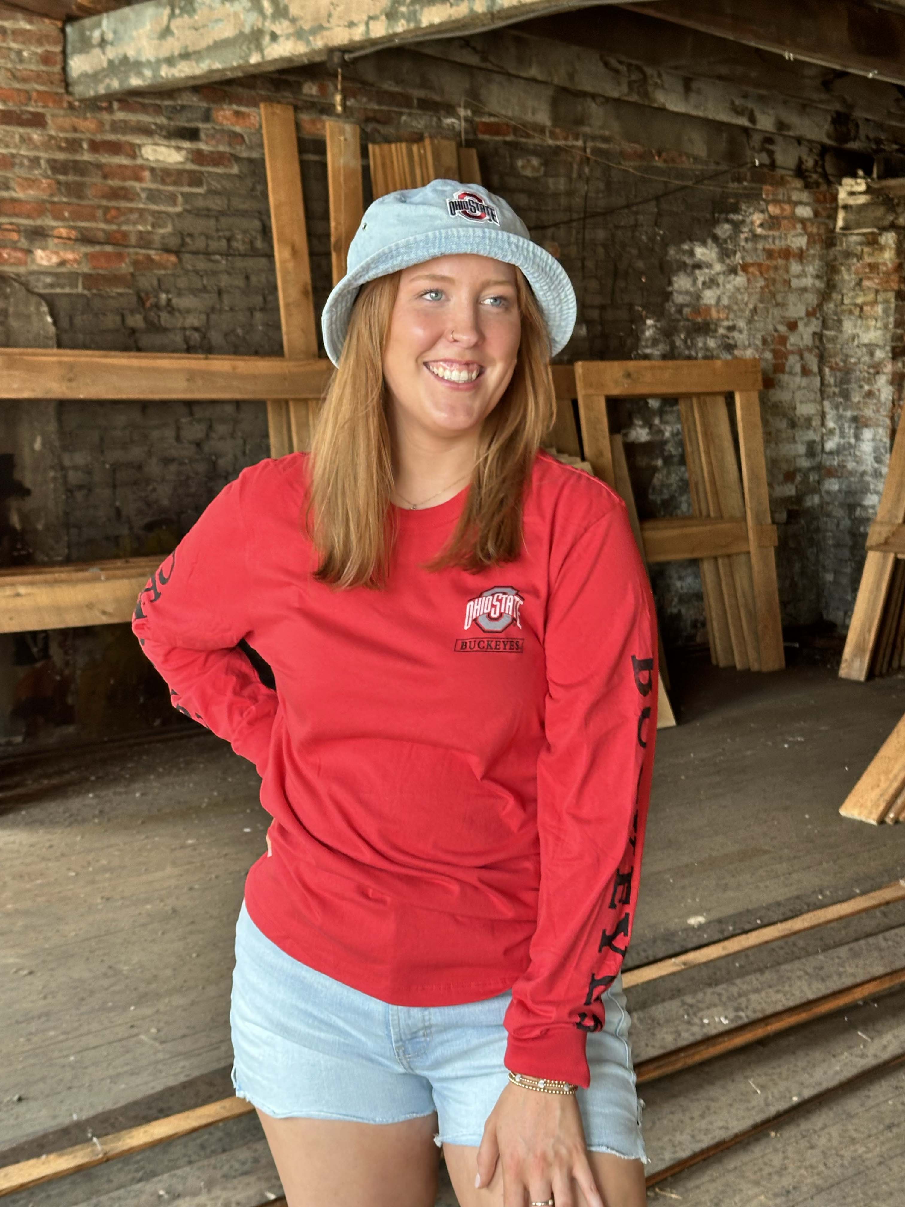 A smiling woman wearing a red long-sleeve t-shirt with a 'Pittsford' logo, a light blue bucket hat, and denim shorts, standing in a rustic workshop with brick and wood walls.