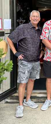 An older man standing outdoors in front of a storefront doorway, wearing a dark patterned short-sleeve polo shirt, gray cargo shorts, and light gray sneakers. Another person is partially visible on the right edge.
