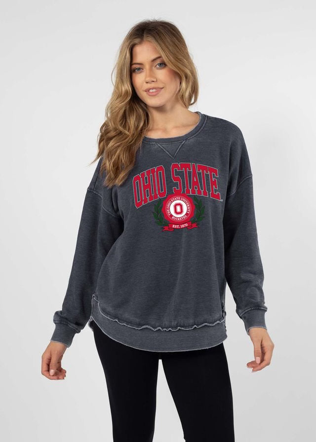 A woman modeling a dark gray washed sweatshirt with a red and white 'Ohio State' graphic on the front, paired with black pants, against a light gray studio background.