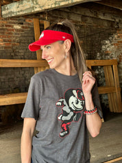 A woman wearing a dark gray graphic t-shirt with a cartoon mascot and a red visor, standing in a rustic brick and wood interior.
