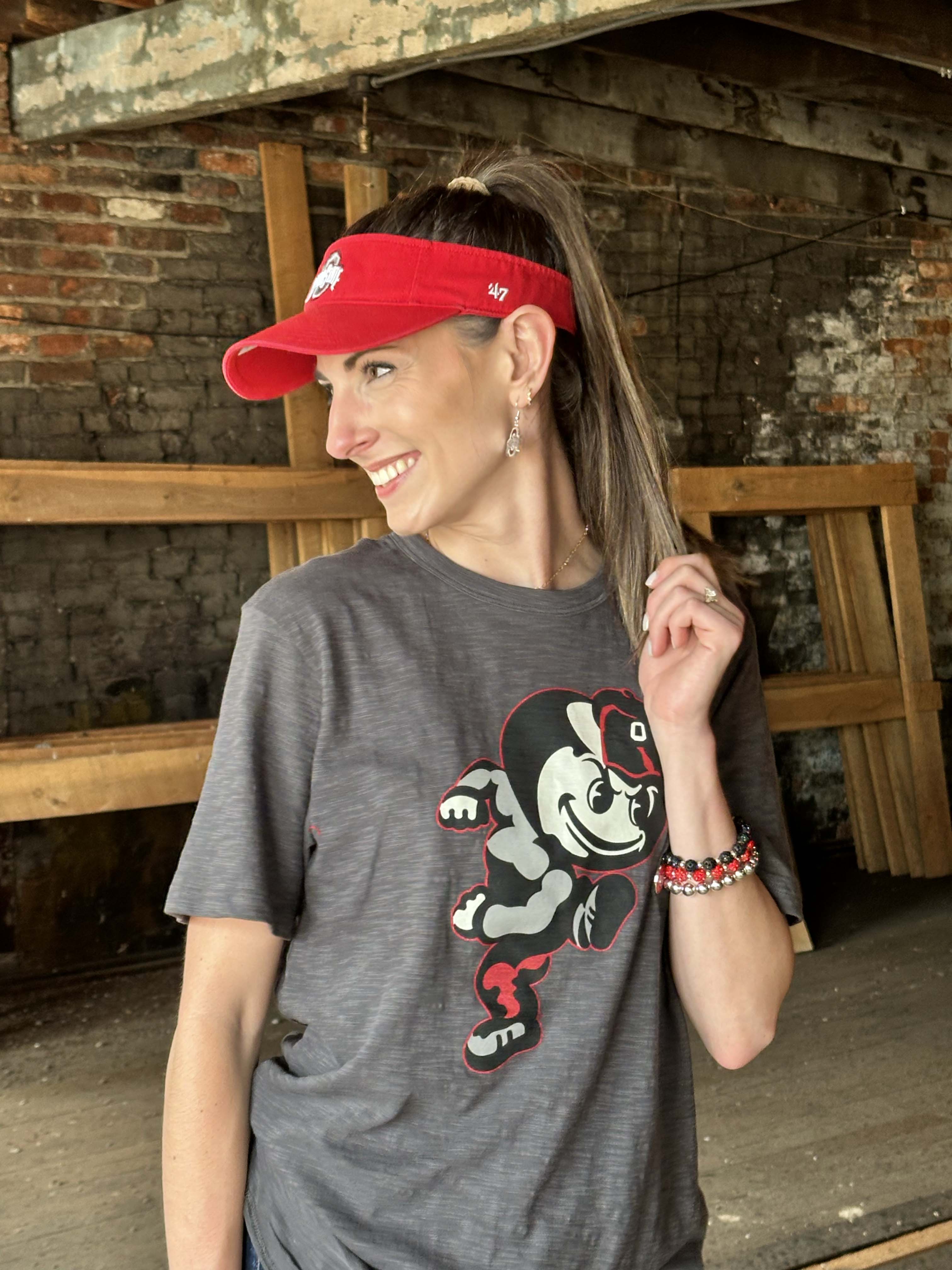 A woman wearing a dark gray graphic t-shirt with a cartoon mascot and a red visor, standing in a rustic brick and wood interior.