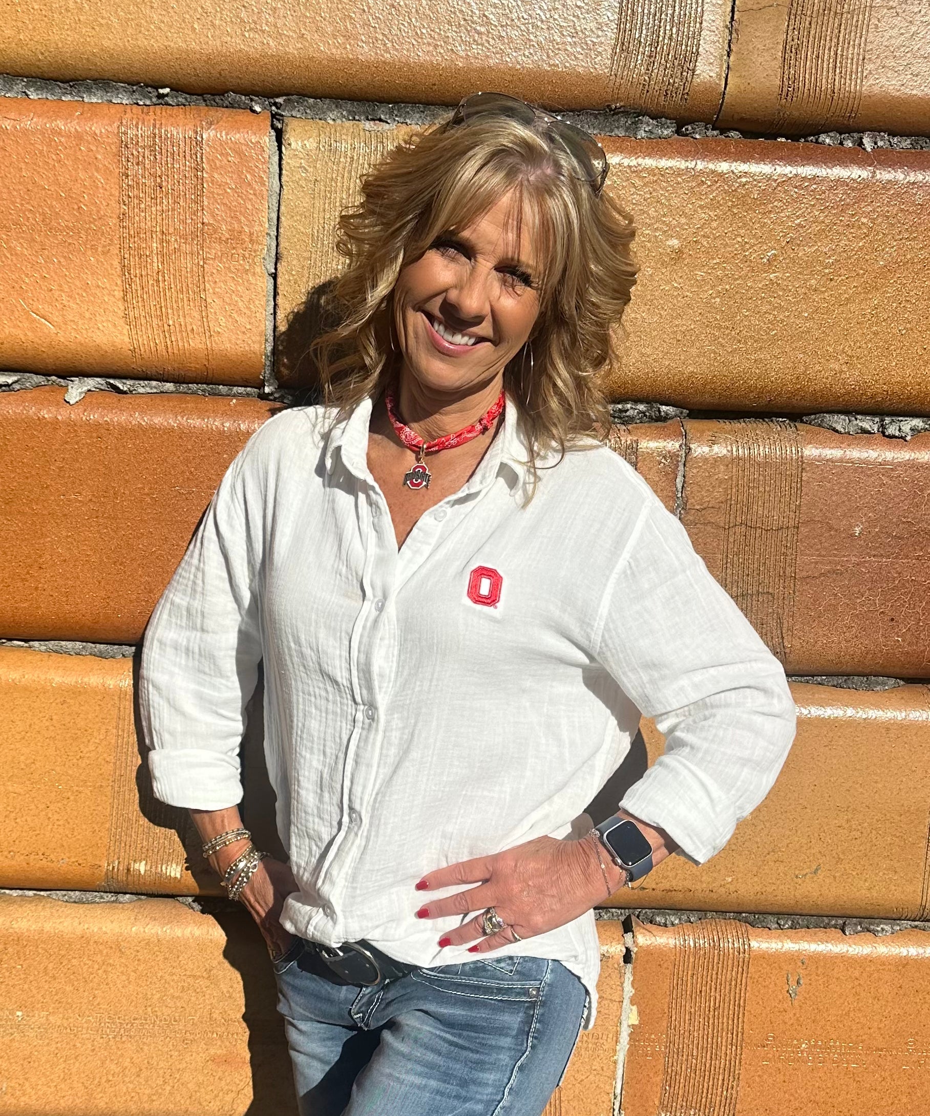A smiling blonde woman wearing a white long-sleeve button-up shirt with a small red logo on the chest, blue jeans, and a red necklace, standing against a tan brick wall.