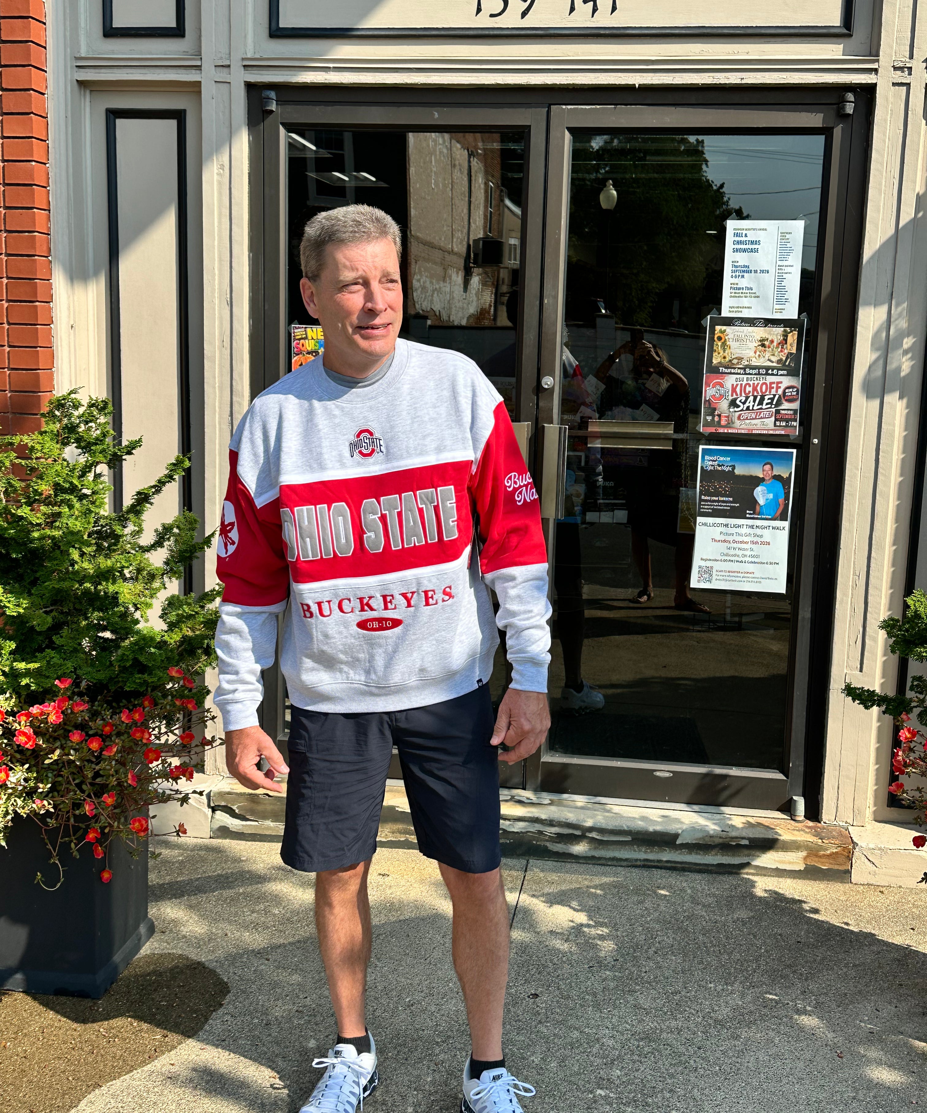 A man wearing an Ohio State Buckeyes long-sleeve sweatshirt, dark shorts, and white sneakers stands in front of a storefront with the address 139 141 on a sunny day.