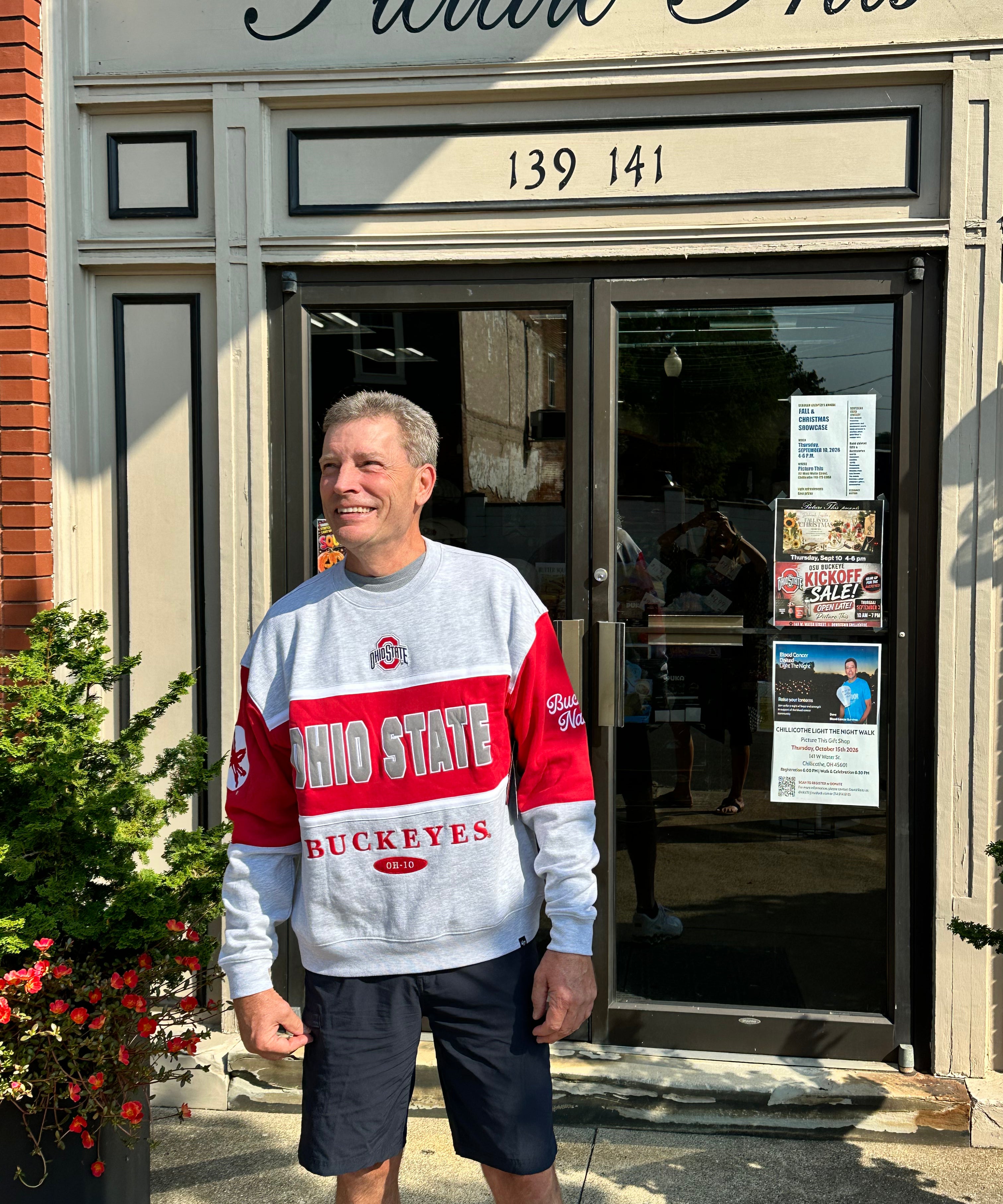 A man wearing a gray and red Ohio State Buckeyes sweatshirt and dark shorts stands in front of a storefront with 'Picture This' written above the door.