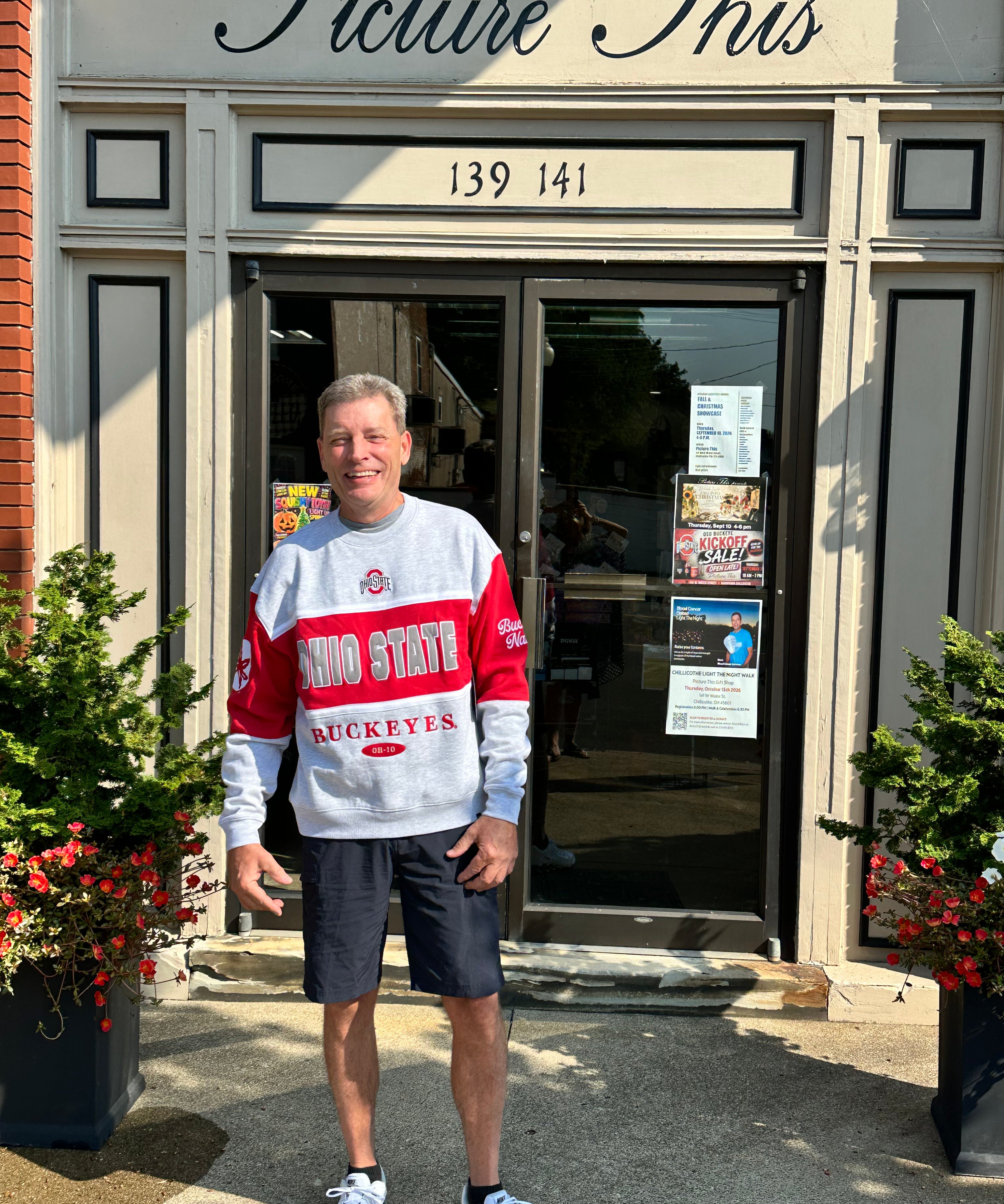 A man stands in front of a storefront wearing a gray and red Ohio State Buckeyes sweatshirt, dark shorts, and white sneakers. The shop has a sign reading 'Picture This' above the entrance.
