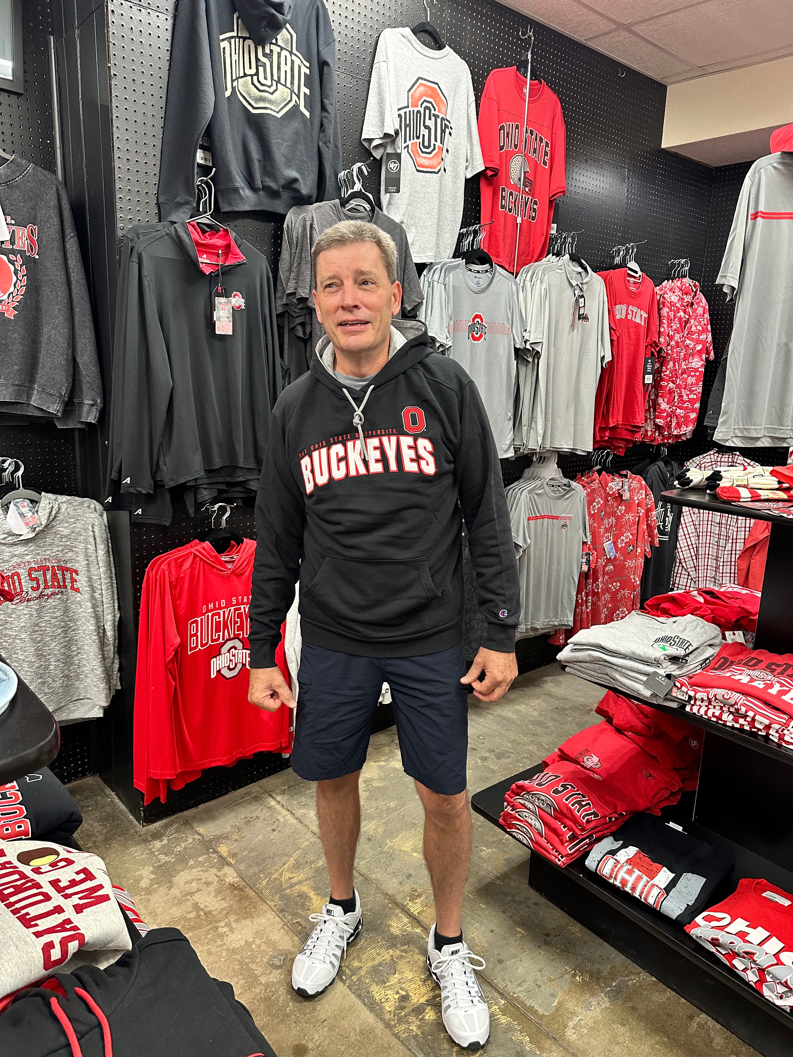 A man wearing a black Ohio State Buckeyes hoodie and navy shorts standing inside a retail store surrounded by Ohio State Buckeyes merchandise.