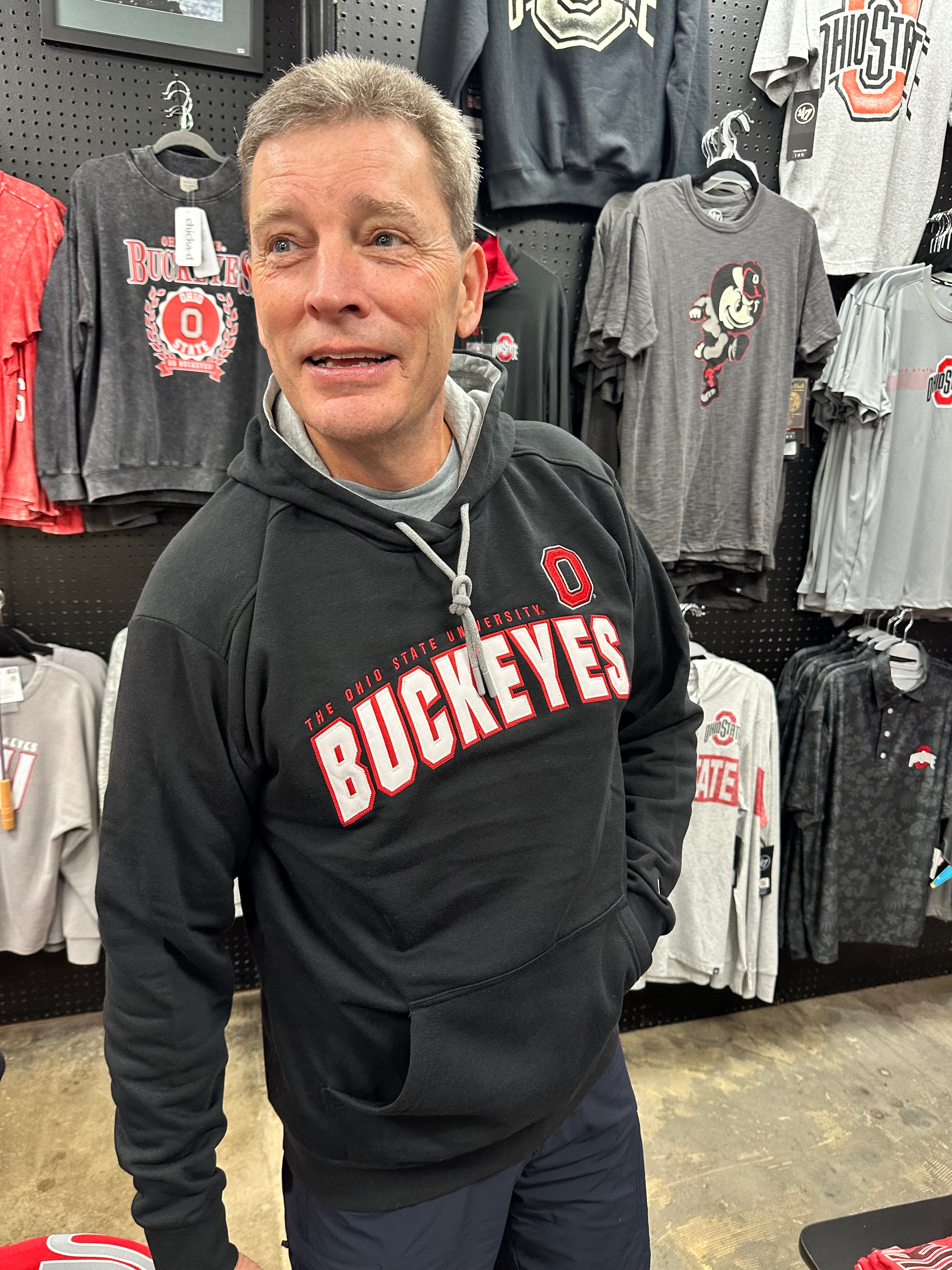 A man wearing a black Ohio State Buckeyes pullover hoodie stands in a retail store, with various Ohio State themed t-shirts hanging on the pegboard behind him.