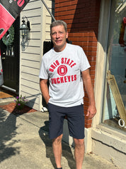 A man standing outdoors on a sidewalk in front of a brick and siding building, wearing a light gray t-shirt with red 'Ohio State Buckeyes' lettering and dark navy shorts.