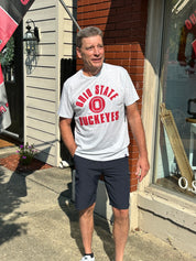 A man wearing a light gray t-shirt with red 'Ohio State Buckeyes' lettering and dark navy shorts, standing outdoors on a sidewalk in front of a brick and siding building.