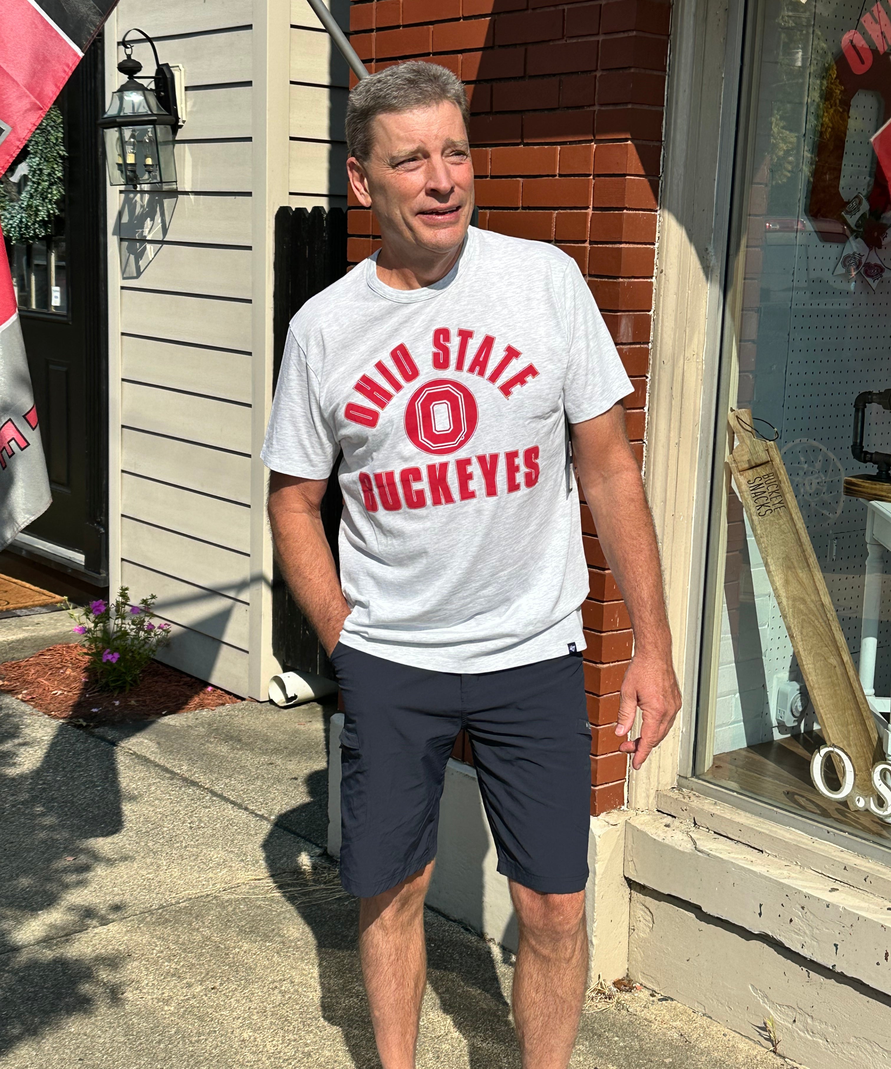 A man wearing a light gray t-shirt with red 'Ohio State Buckeyes' lettering and dark navy shorts, standing outdoors on a sidewalk in front of a brick and siding building.