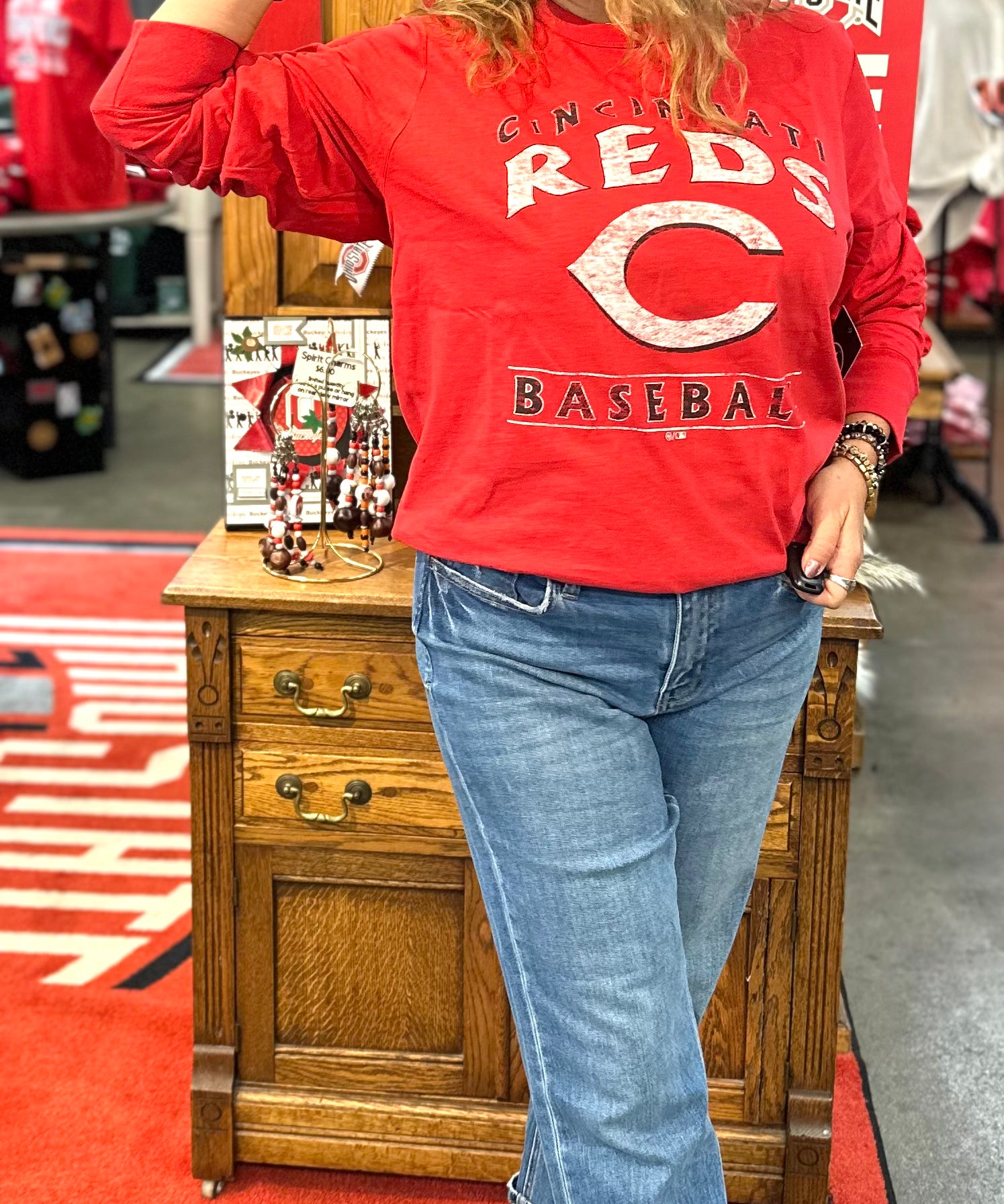 Person wearing a red 'Reds Baseball' sweatshirt and blue jeans, standing in a store with promotional signs.