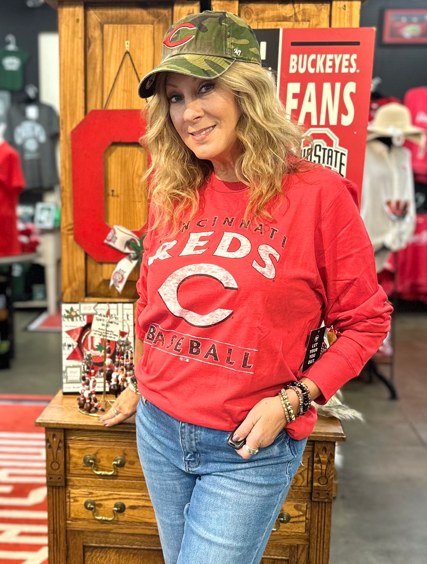 Woman wearing a red 'Reds Baseball' sweatshirt and green cap in a store setting.