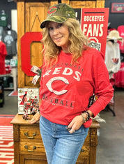 Woman wearing a red 'Reds Baseball' sweatshirt and green cap in a store setting.