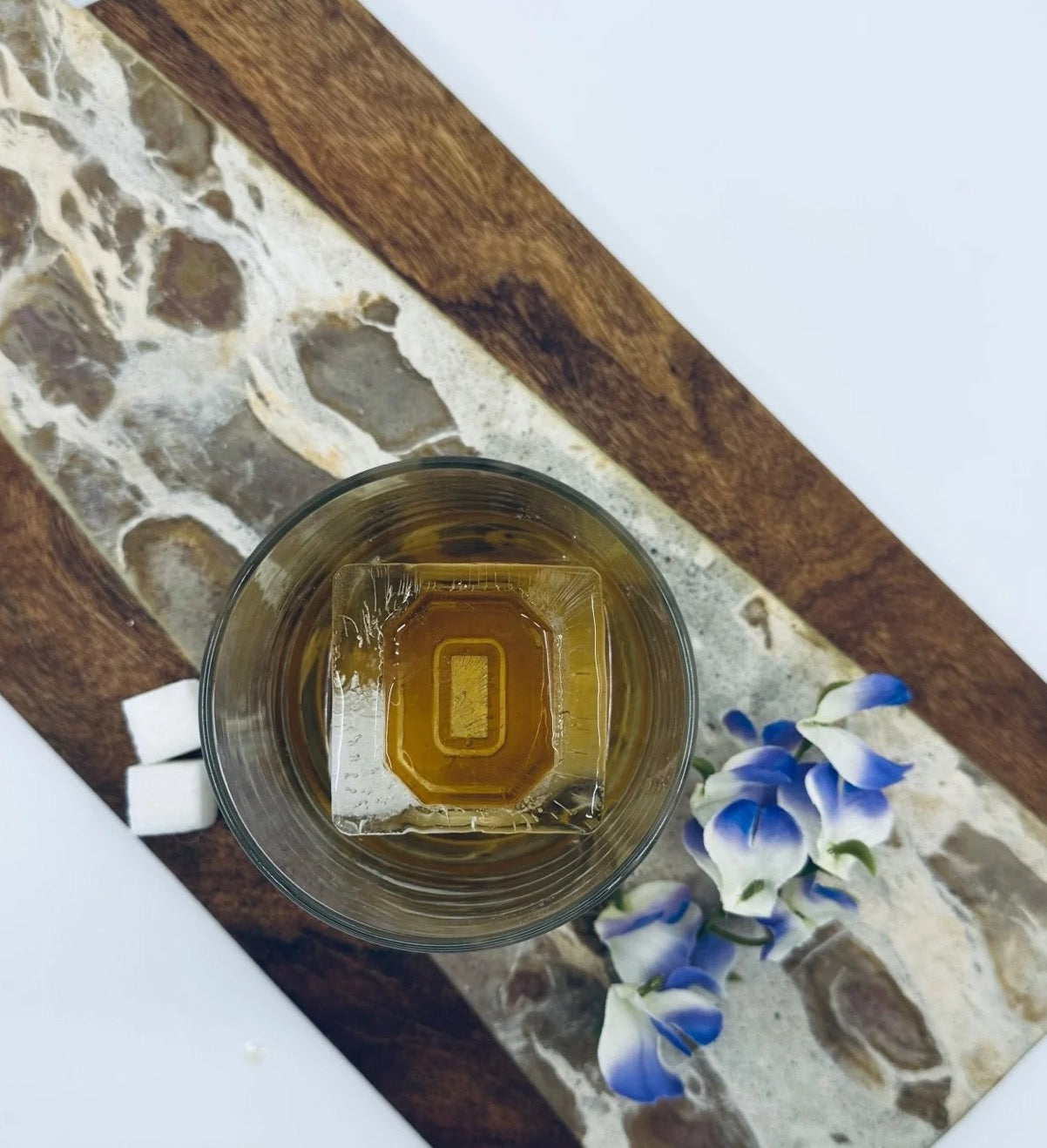 Glass with a square ice cube and liquid on a stone and wood surface with flowers.
