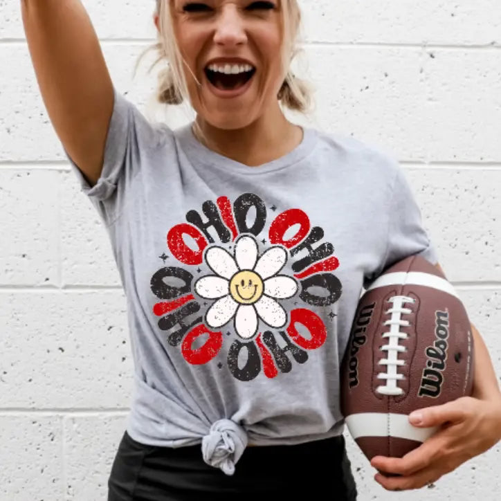 Person wearing a gray t-shirt with a colorful flower design, holding a football against a white brick wall.