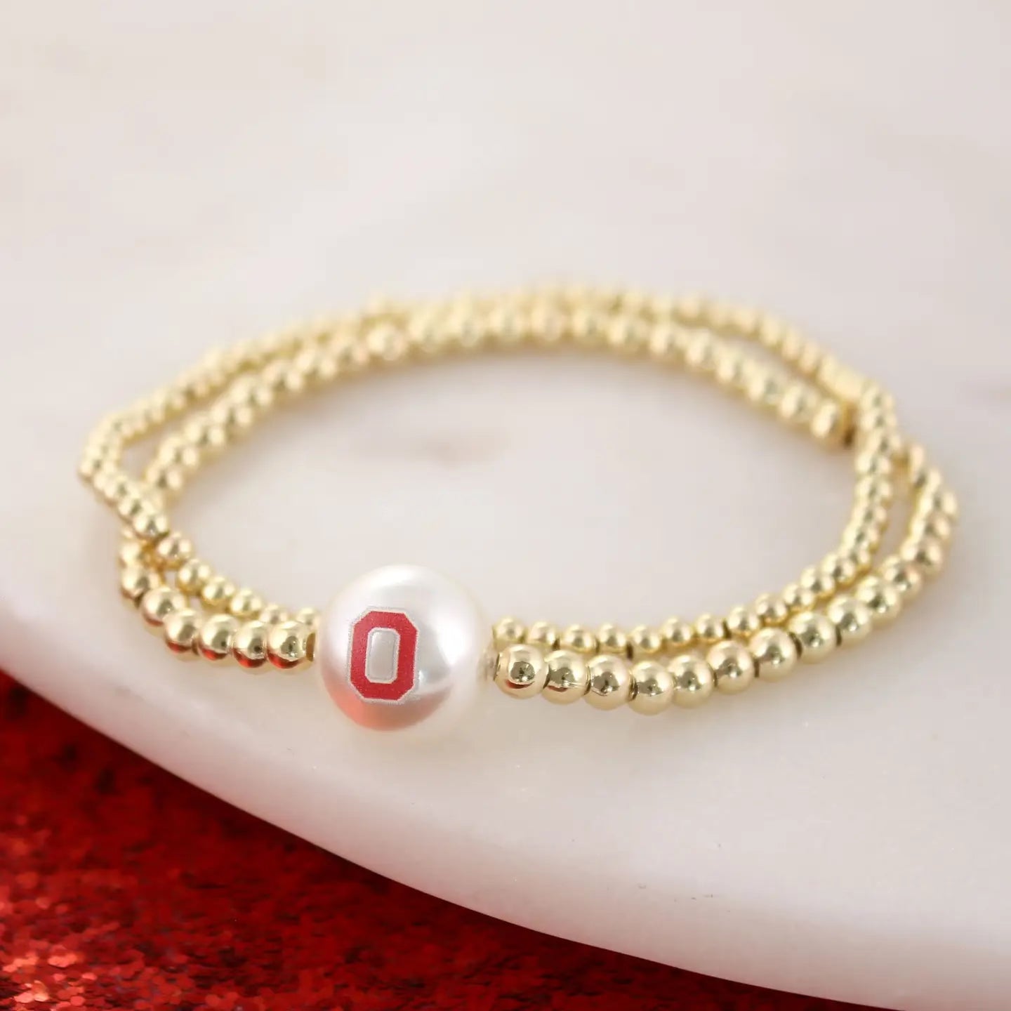 A gold beaded double-strand bracelet featuring a white pearl charm with a red letter 'O' emblem, resting on a white marble surface with a red glittery surface in the background.