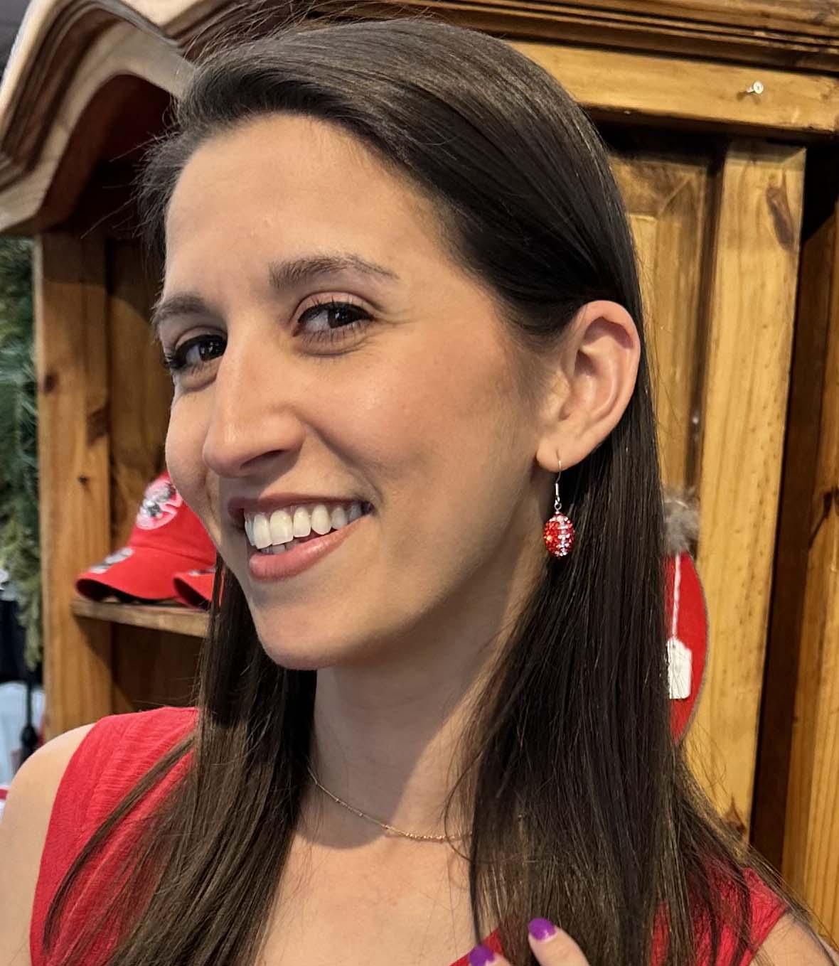 A smiling woman with long dark brown hair wearing a red top and a small red dangling earring with a white letter 'M' and a red heart, standing in front of a wooden display cabinet.